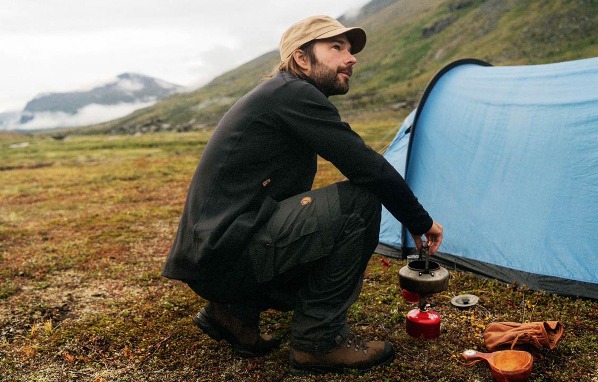 a man crouched by a tent using a camp stove in Fjallraven apparel