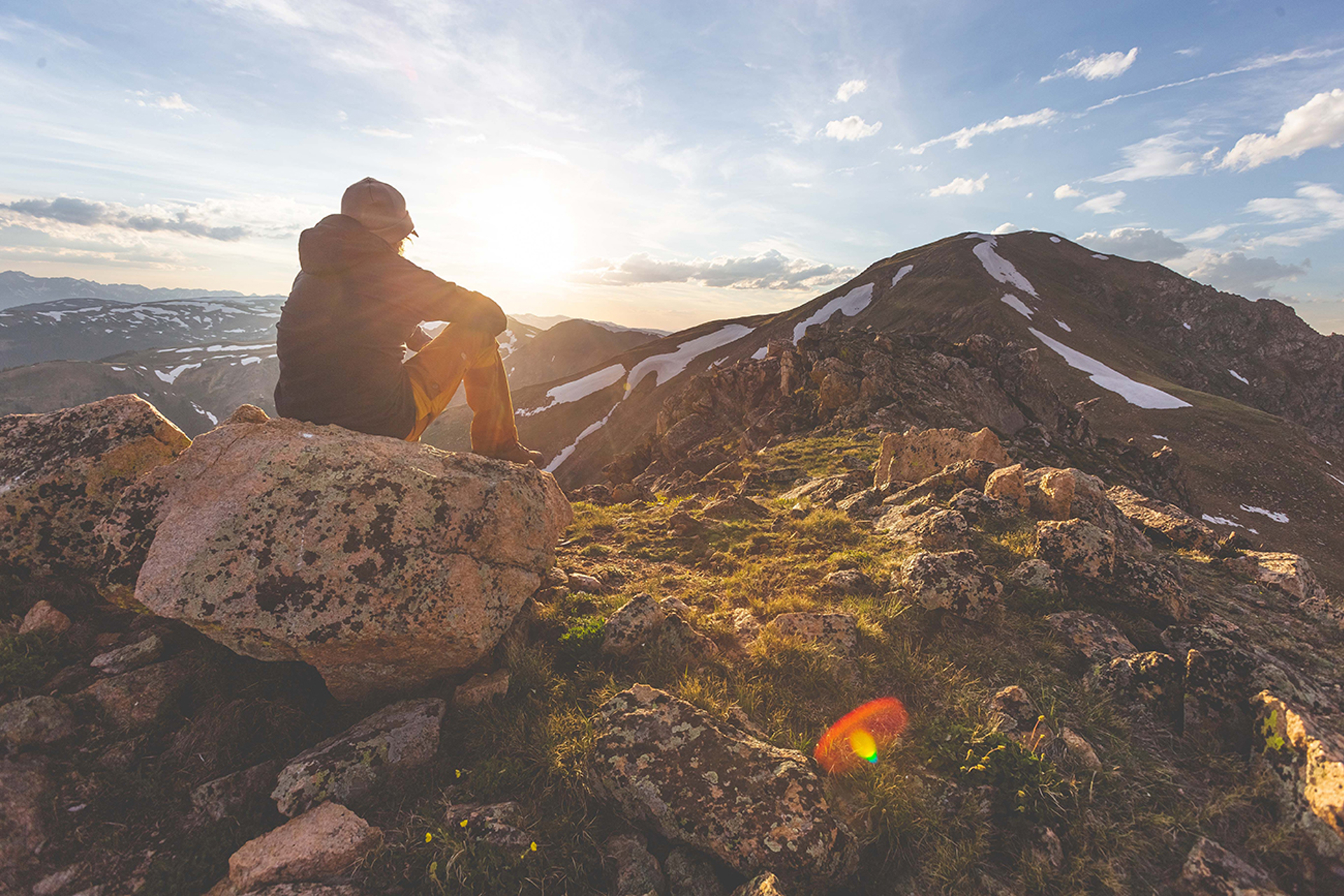 Man in hat, jacket, and yellow pants sitting on rocky mountain top looking over other mountain tops at sunset.
