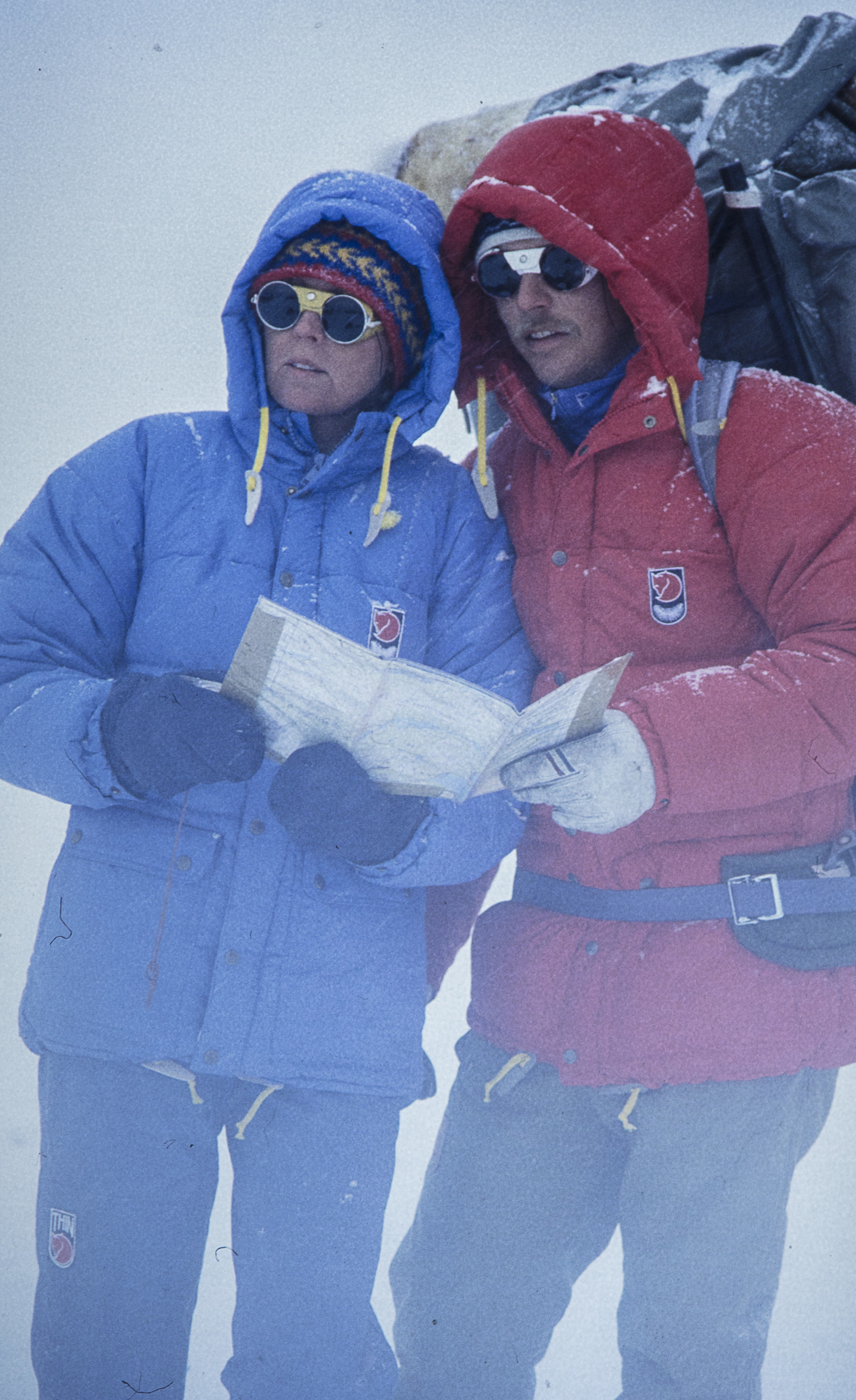 Man and woman in heavy Fjallraven parkas standing together in snow storm reviewing a map with hoods, gloves, and sunglasses on.  