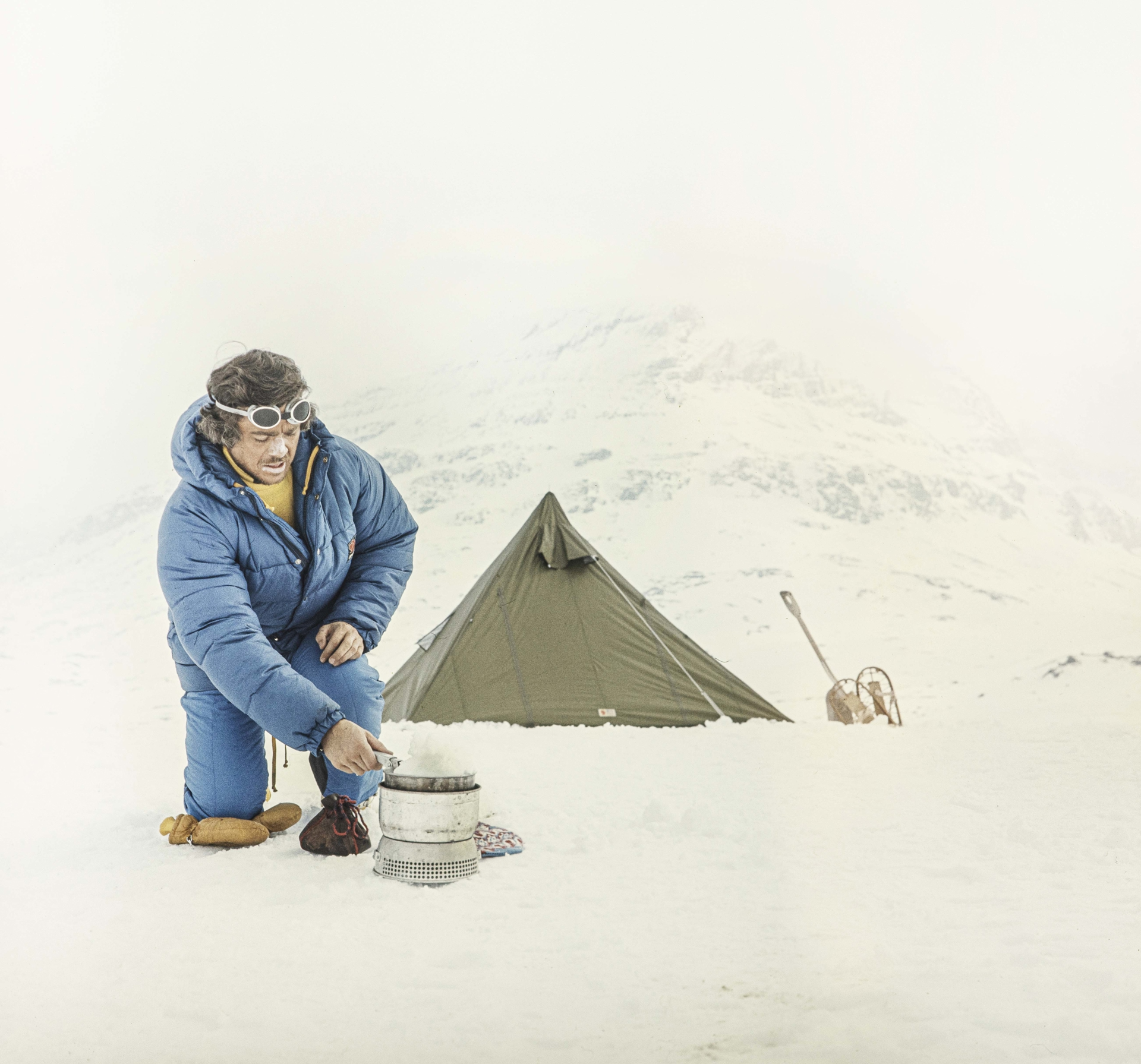 Old picture of man kneeling in blue Fjallraven parka on snow with small green tent in background.
